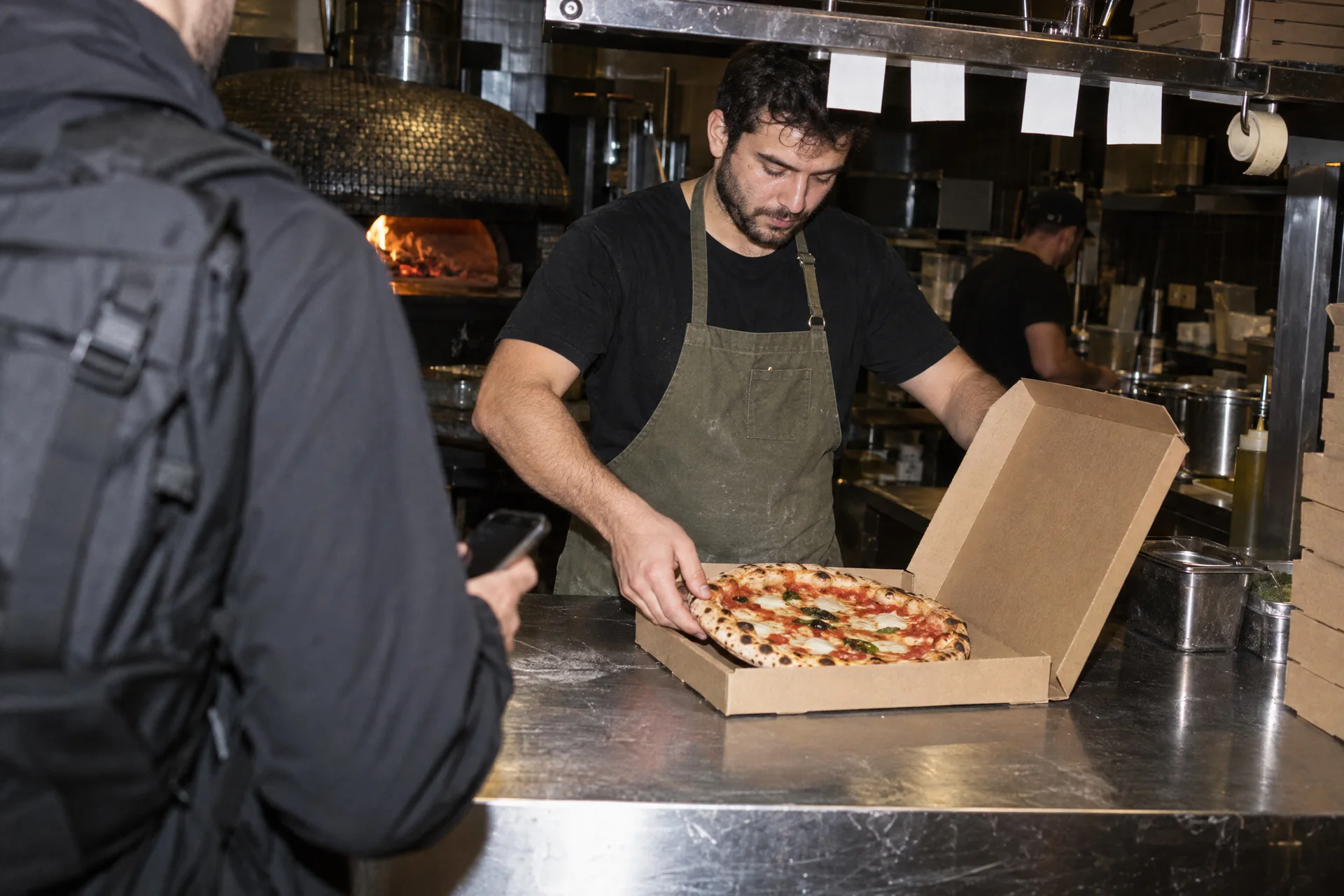 A pizza maker puts a pizza in its box while a courier waits with his phone.